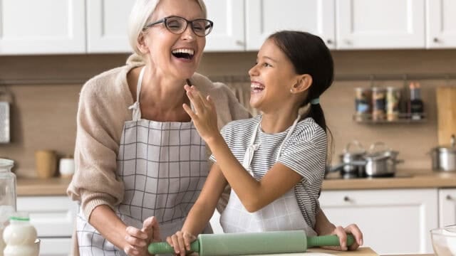An older woman and a young girl, both wearing aprons, laugh together in a kitchen while baking. The girl holds a rolling pin, and they seem to be enjoying their time together.