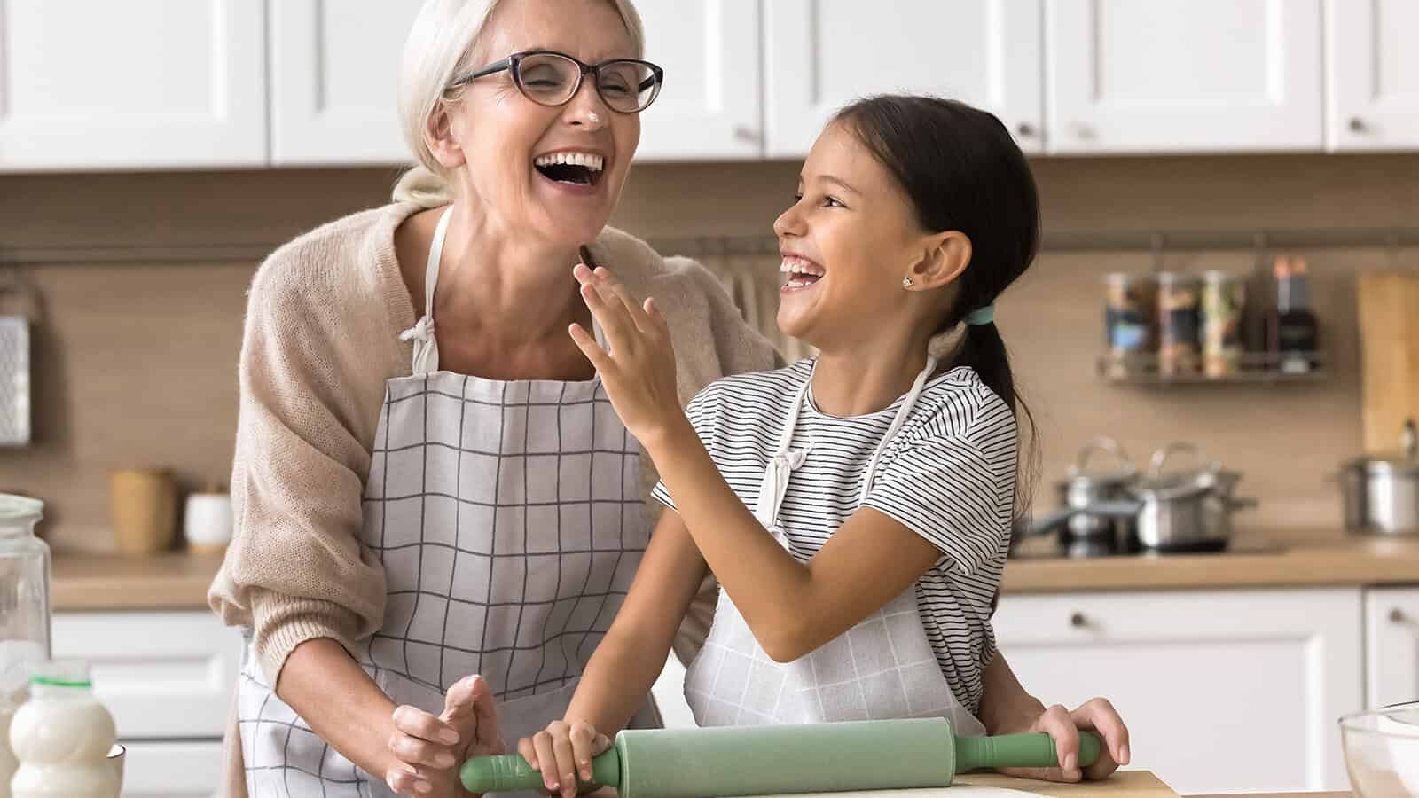 An older woman and a young girl, both wearing aprons, laugh together in a kitchen while baking. The girl holds a rolling pin, and they seem to be enjoying their time together.