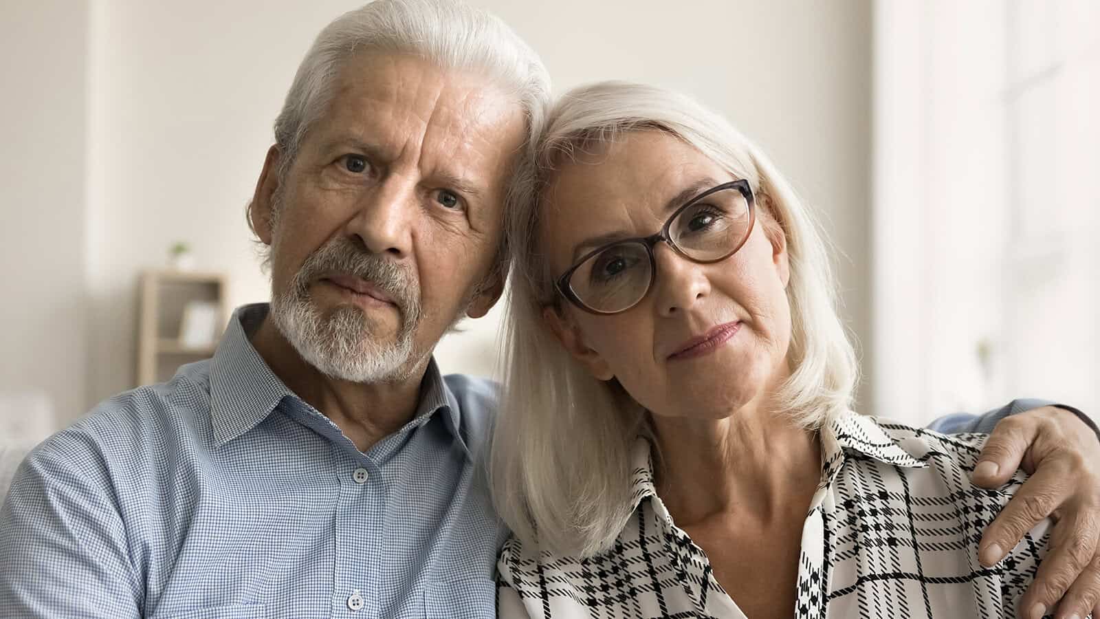 An older man and woman sit close together, looking at the camera. The man wears a light blue shirt and the woman has glasses and a patterned blouse. They both have gray hair and calm, affectionate expressions.