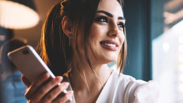 A woman with long dark hair holds a smartphone and smiles while looking to the side, seated indoors near a window with soft lighting.