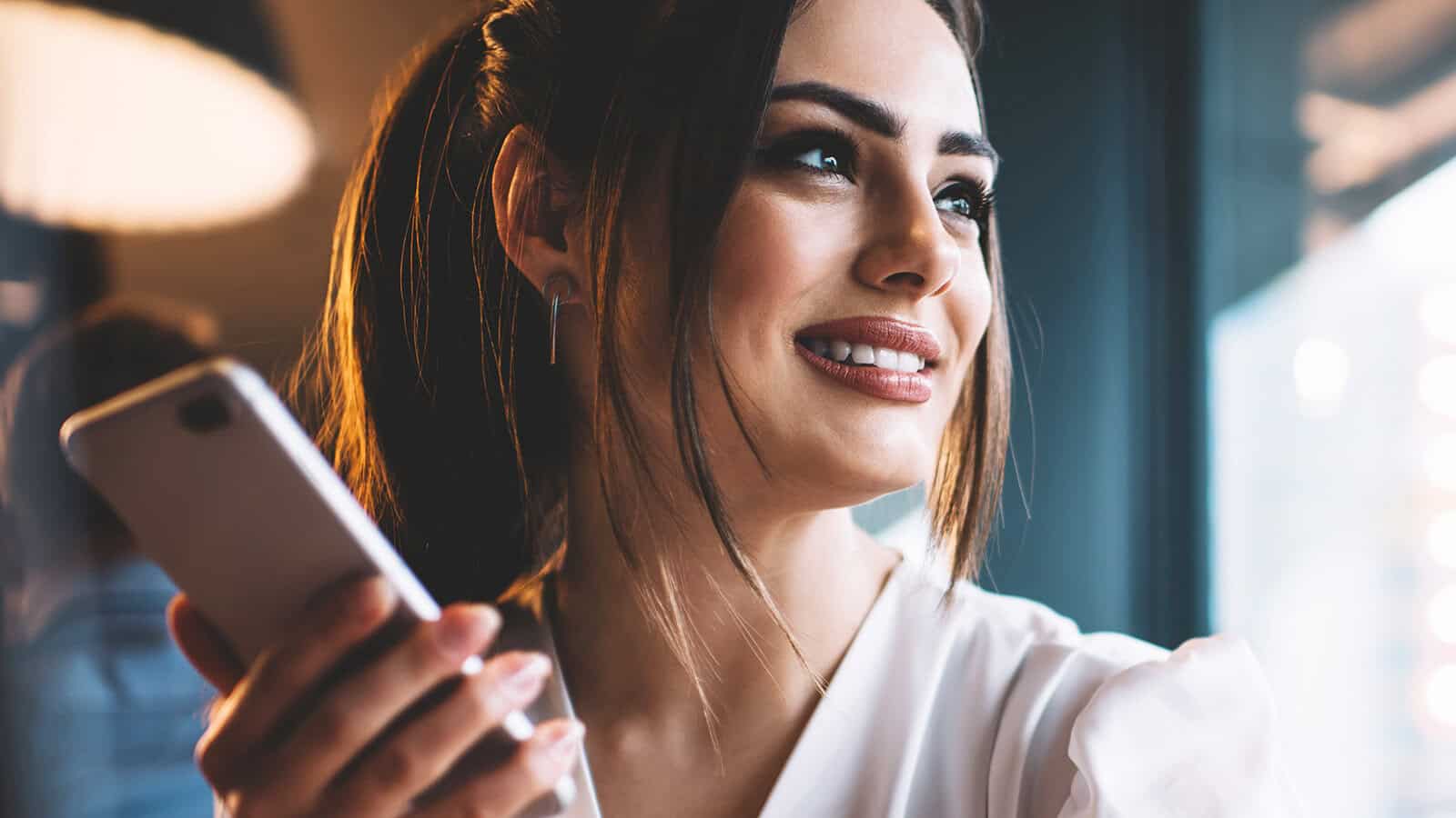 A woman with long dark hair holds a smartphone and smiles while looking to the side, seated indoors near a window with soft lighting.