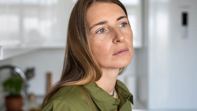 A woman with long brown hair, wearing a green shirt, looks thoughtful while standing in a bright kitchen.