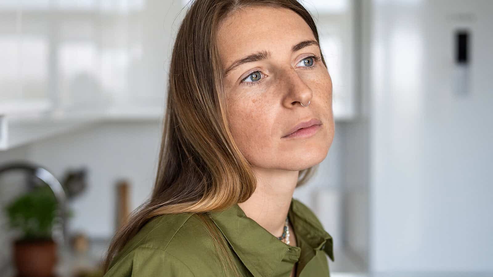 A woman with long brown hair, wearing a green shirt, looks thoughtful while standing in a bright kitchen.