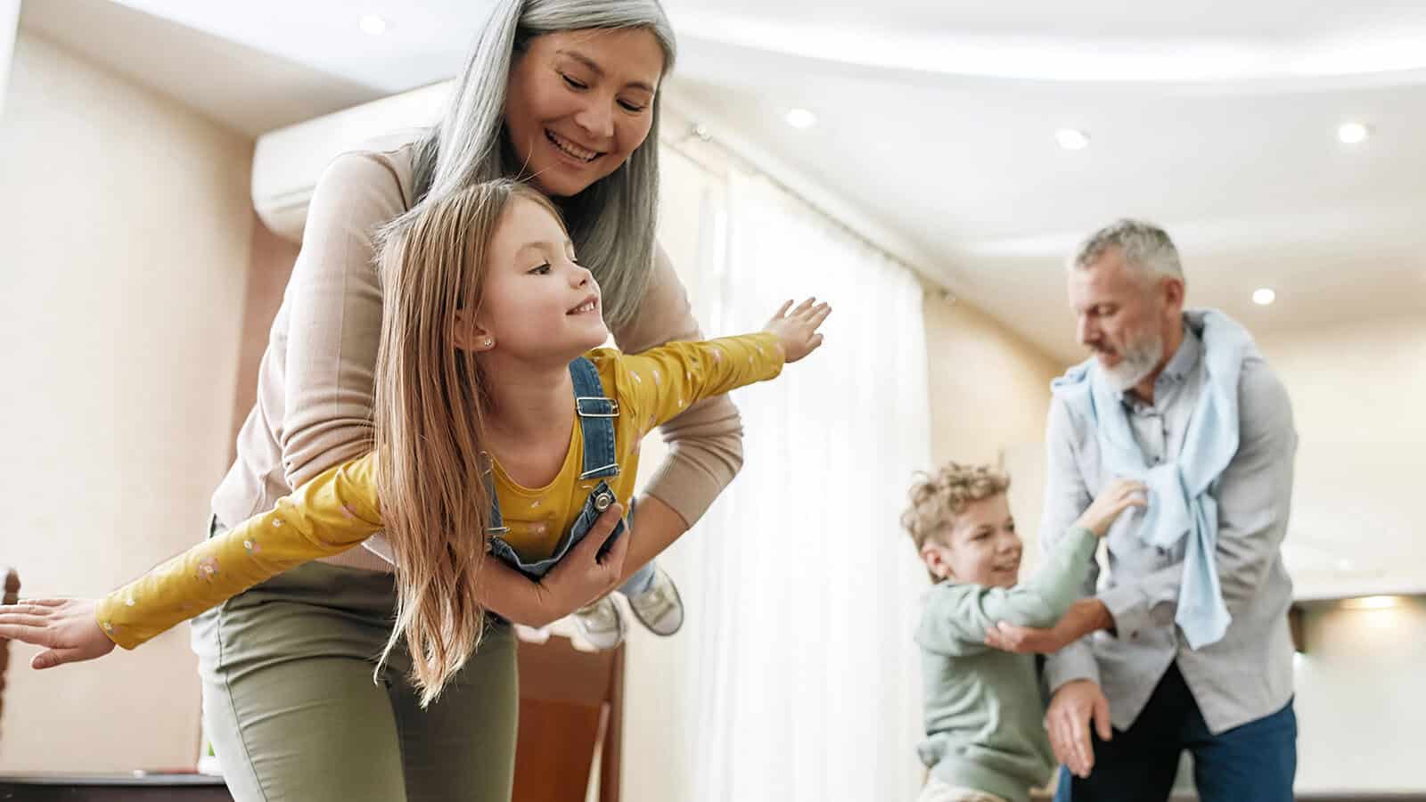 A woman holds a smiling girl with outstretched arms like an airplane, while a man plays with a laughing boy in a bright living room.