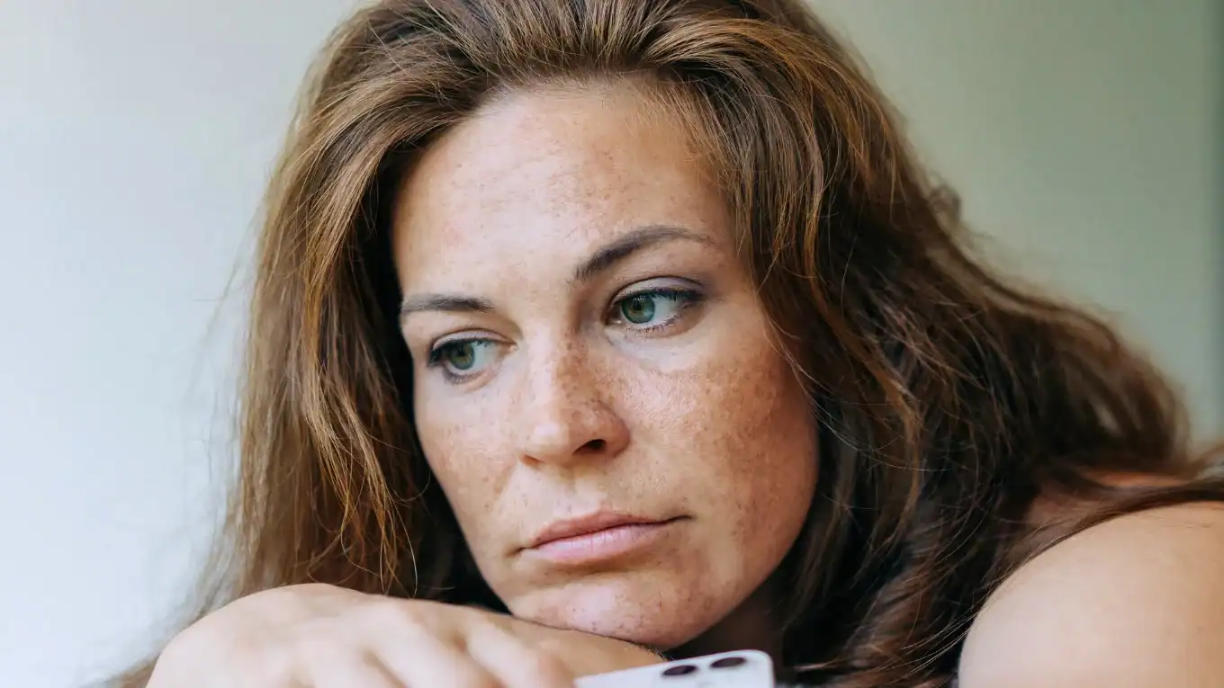 A woman with long brown hair and freckles looks down with a sad or thoughtful expression, resting her chin on her hand while holding a smartphone.