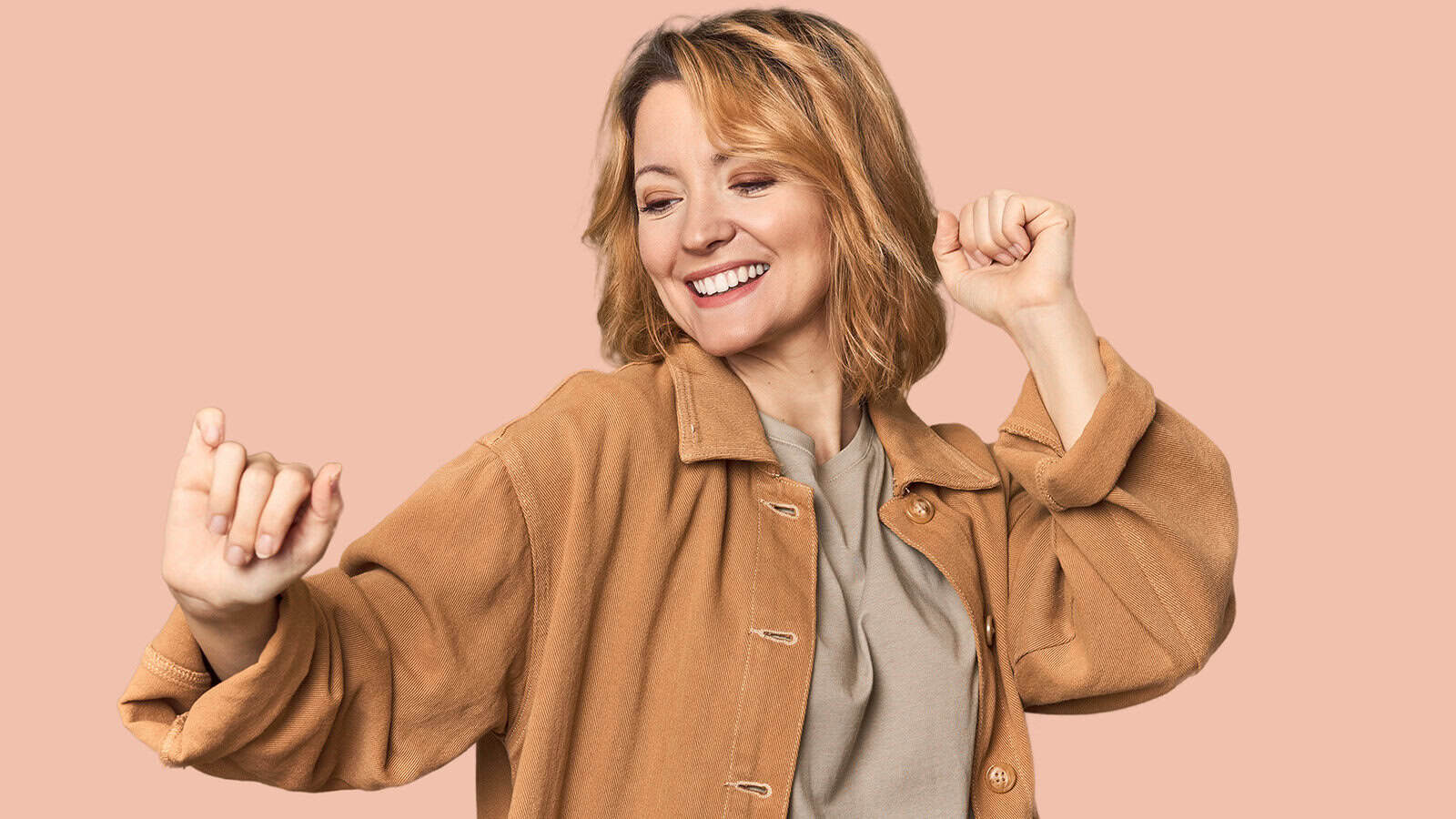 A woman with short blond hair smiles and dances, wearing a tan jacket over a beige shirt, in front of a solid peach background—a joyful moment that hints at leaving parenting guilt behind and embracing new beginnings, much like the spirit of back to school.