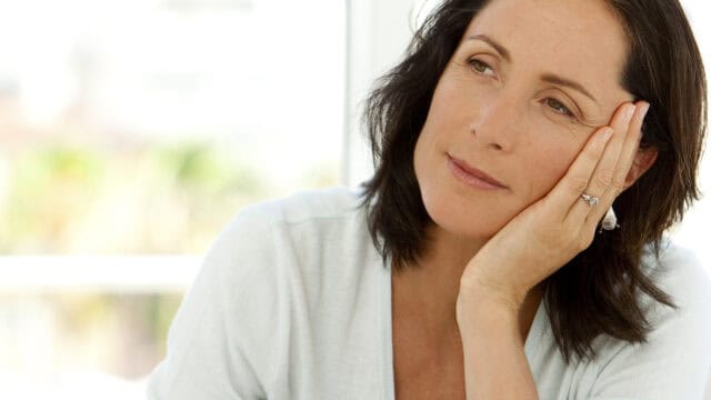 A woman with dark hair rests her head on her hand, gazing thoughtfully into the distance. She wears a light-colored top and a ring on her finger, and appears calm and reflective.