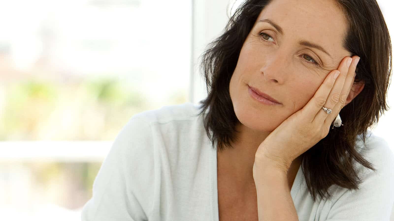 A woman with dark hair rests her head on her hand, gazing thoughtfully into the distance. She wears a light-colored top and a ring on her finger, and appears calm and reflective.