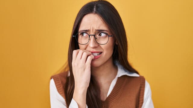 A young woman with glasses looking anxious as she bites her fingernails.
