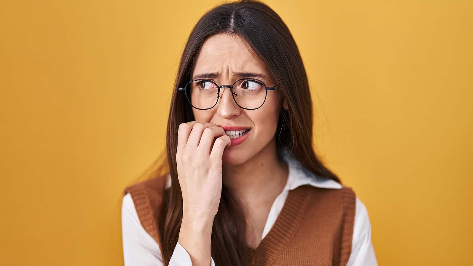 A young woman with glasses looking anxious as she bites her fingernails.