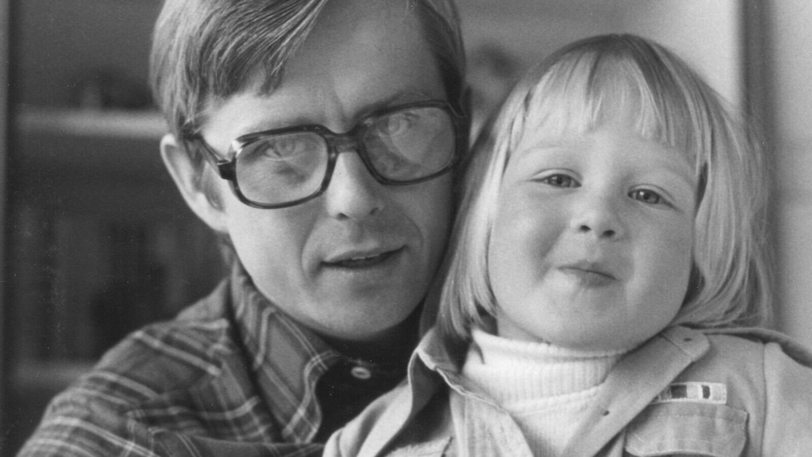 A black and white close-up portrait of a man wearing glasses and a young child leaning against his shoulder.
