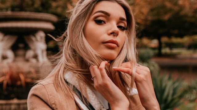 A woman with long blonde hair and a nose ring poses outdoors near a stone fountain, touching her hair. She wears a tan jacket with a white collar. Blurred greenery and autumn colors are in the background.