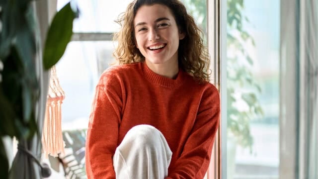 A smiling woman with curly hair wearing an orange sweater sits by a window.