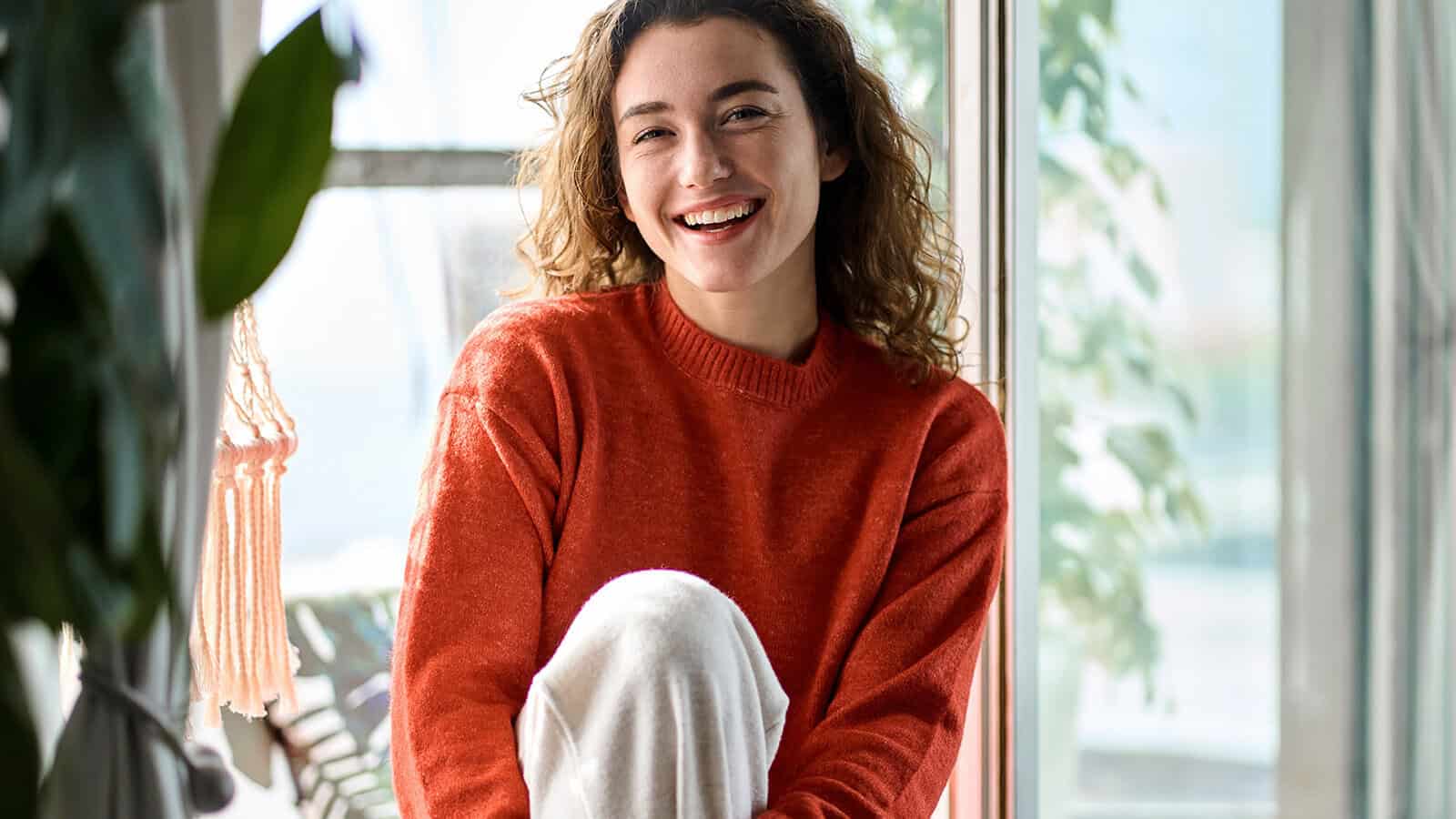 A smiling woman with curly hair wearing an orange sweater sits by a window.