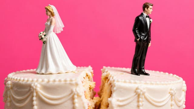 A wedding cake topper featuring a bride and groom figurine standing on separate halves of a sliced cake against a pink background.