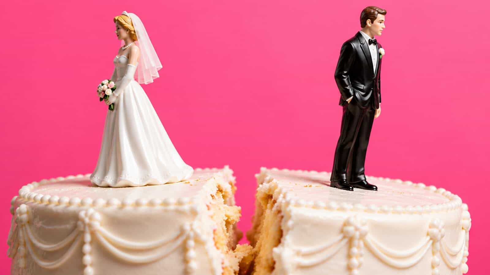A wedding cake topper featuring a bride and groom figurine standing on separate halves of a sliced cake against a pink background.