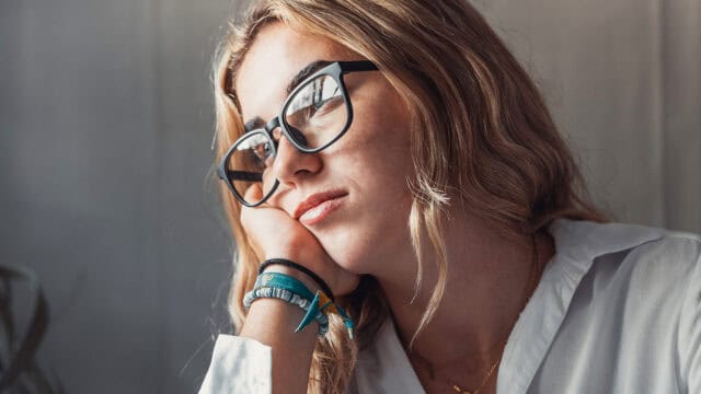 A young woman with glasses rests her chin on her hand, looking away with a thoughtful expression.