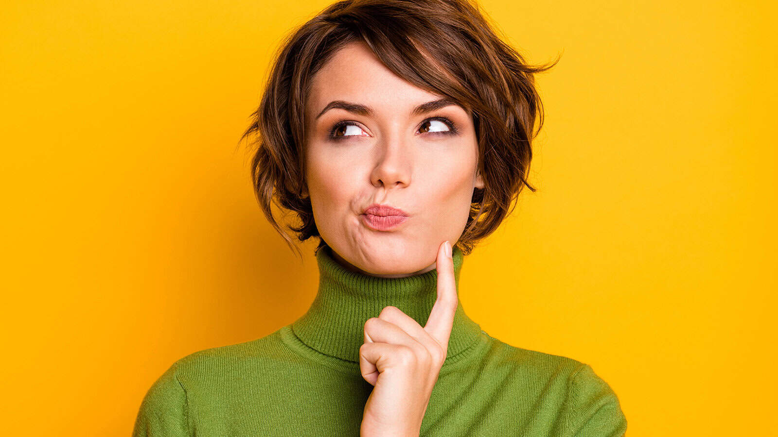 A person in a green turtleneck sweater looking thoughtful with a hand to their chin.