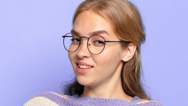 A young woman wearing thin-rimmed, round black glasses looks toward the camera with a slight smile against a light purple background.