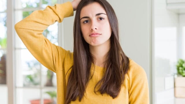 A young woman in a yellow sweatshirt looking confused while scratching her head.