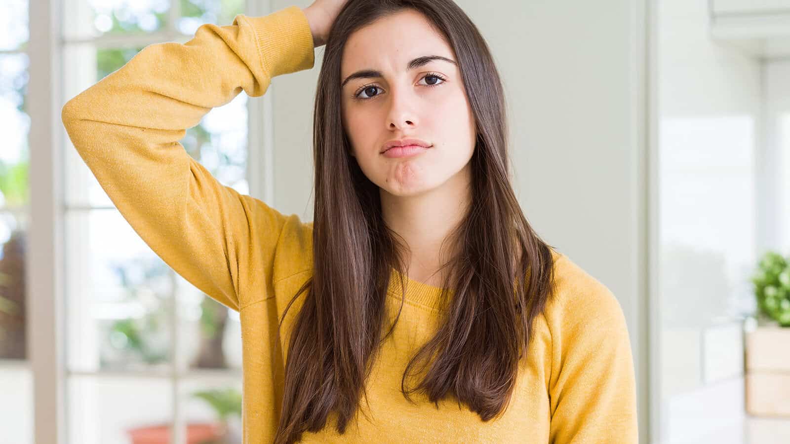 A young woman in a yellow sweatshirt looking confused while scratching her head.
