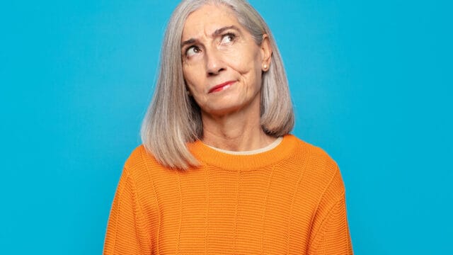 A woman with grey hair wearing an orange sweater looks upward with a thoughtful or skeptical expression.