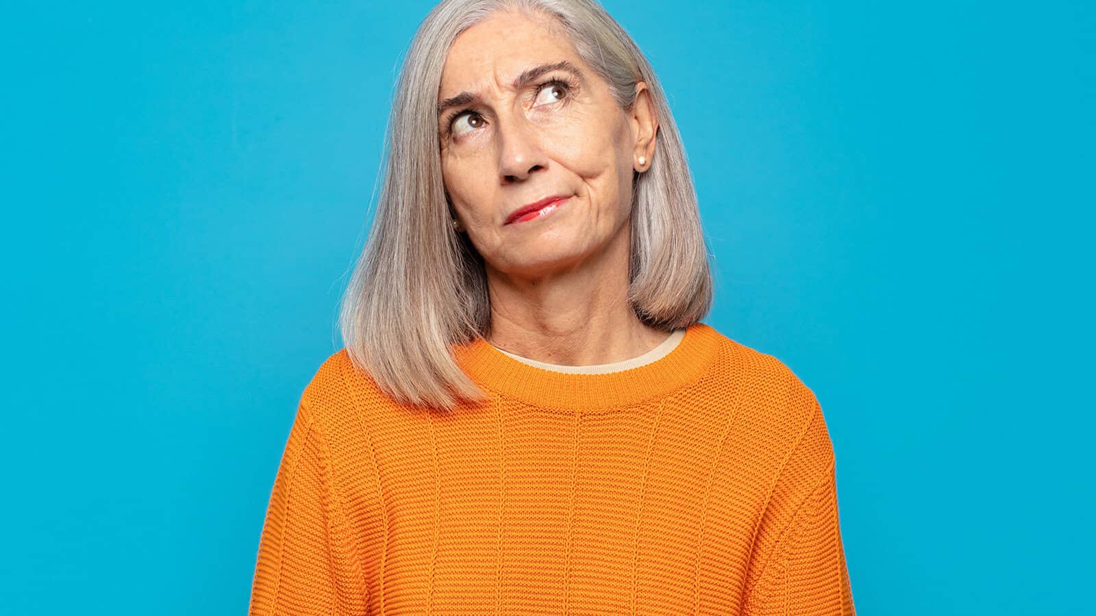 A woman with grey hair wearing an orange sweater looks upward with a thoughtful or skeptical expression.
