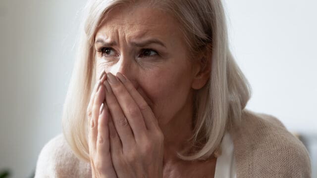 An older woman with gray hair looks worried, covering her mouth and nose with both hands, with a distressed expression on her face.
