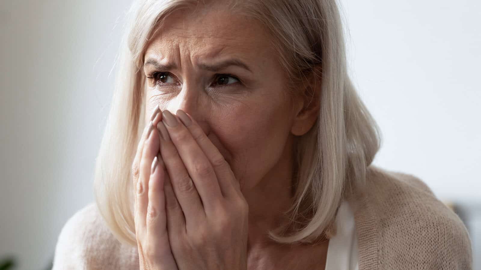 An older woman with gray hair looks worried, covering her mouth and nose with both hands, with a distressed expression on her face.