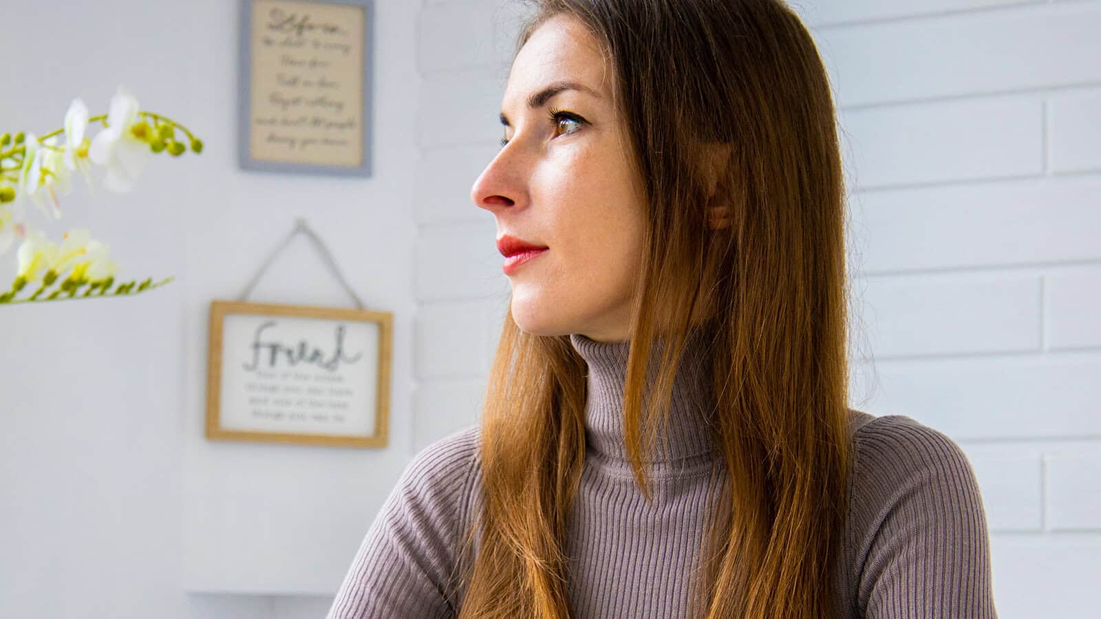 A woman with long brown hair wearing a gray turtleneck looks toward the left, with framed wall art and white flowers visible in the background.