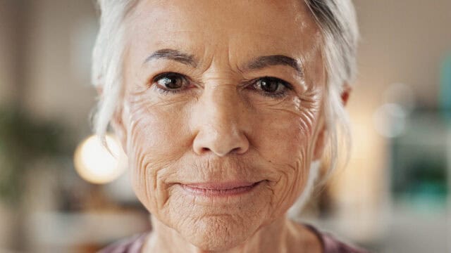 Close-up of an older woman with gray hair and a slight smile, showing facial wrinkles and wearing a mauve shirt. The background is softly blurred.