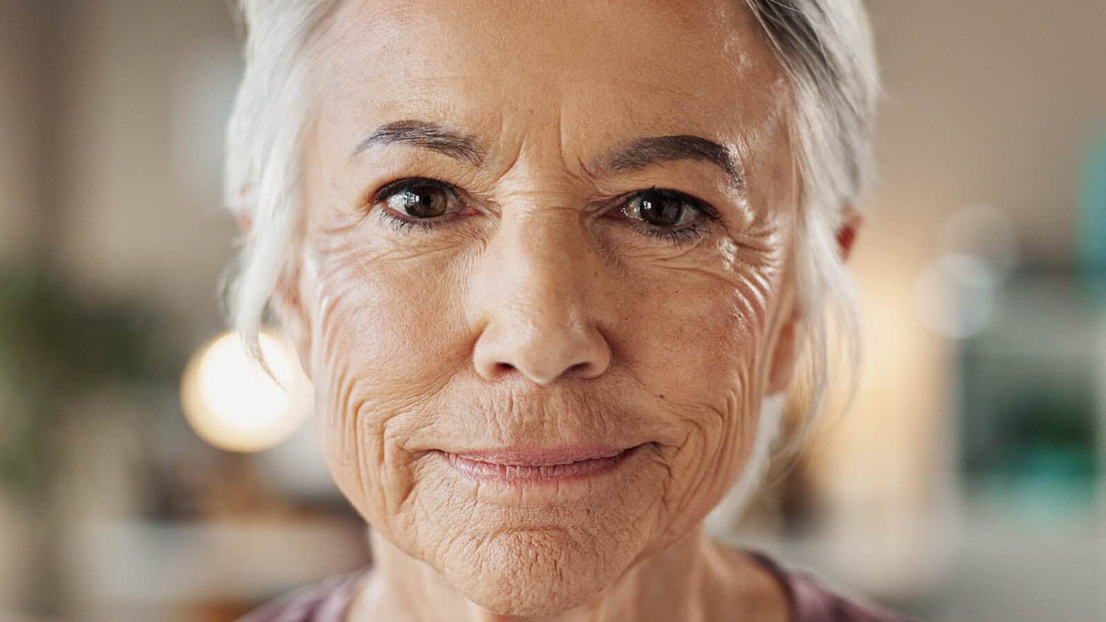 Close-up of an older woman with gray hair and a slight smile, showing facial wrinkles and wearing a mauve shirt. The background is softly blurred.