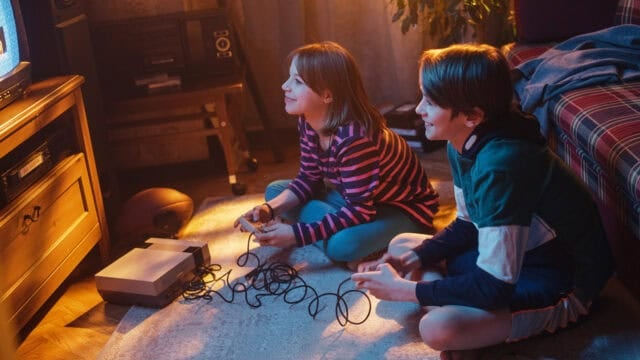 A young girl and boy sit on a rug playing a retro video game console connected to a television.