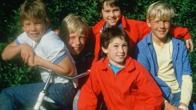 Five boys smiling together outdoors in front of a hedge.