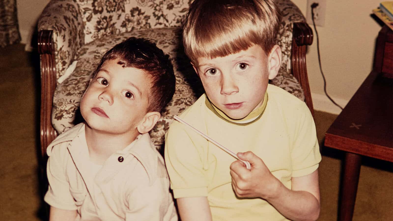 Two young boys sitting side by side, with one holding a drinking straw.