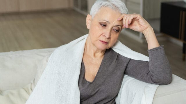 A woman with short white hair rests her hand on her forehead while sitting with a white blanket draped over her shoulders.