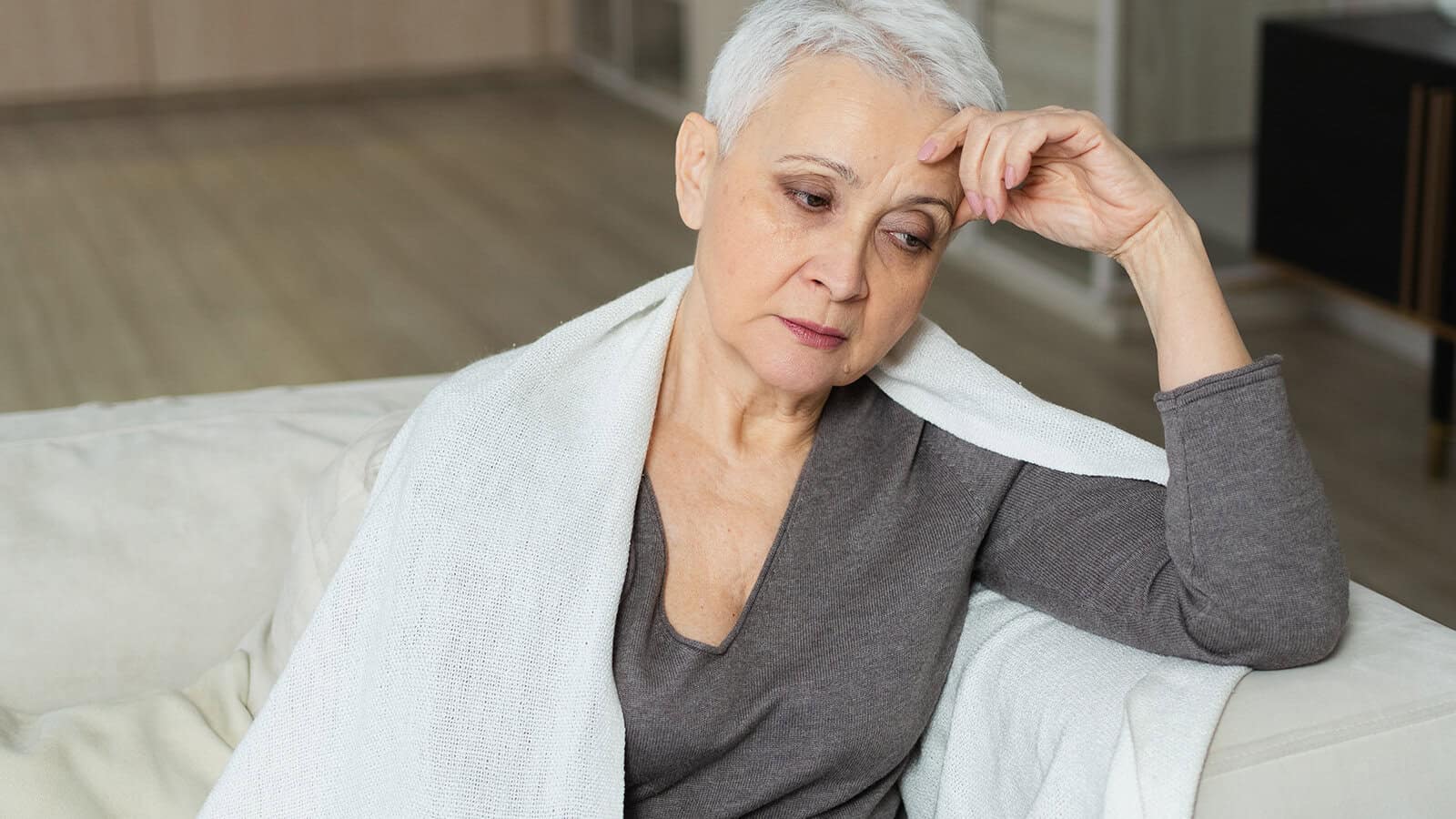 A woman with short white hair rests her hand on her forehead while sitting with a white blanket draped over her shoulders.