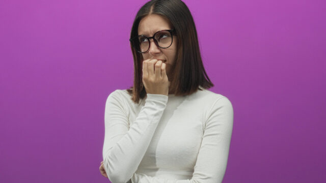 A woman with dark hair and glasses looking sideways while biting her nails in a nervous expression against a purple background.