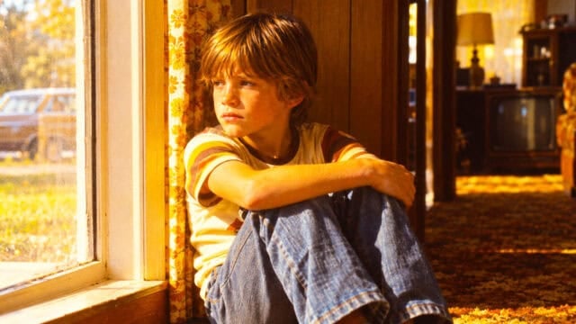 A young boy in a striped t-shirt sits on the floor by a window, looking outside.