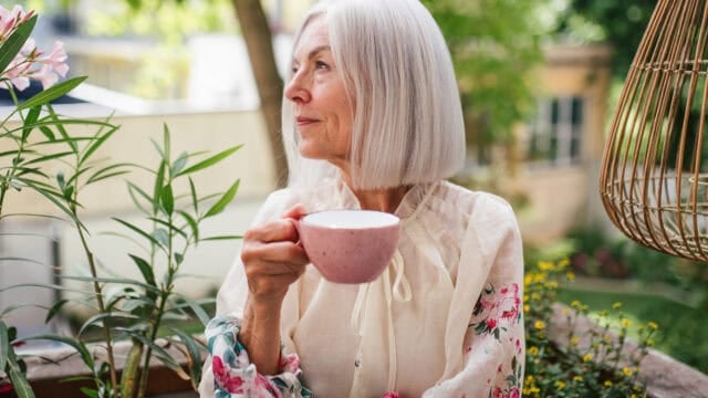 An older woman with shoulder-length grey hair holds a pink mug while looking out over a garden terrace.