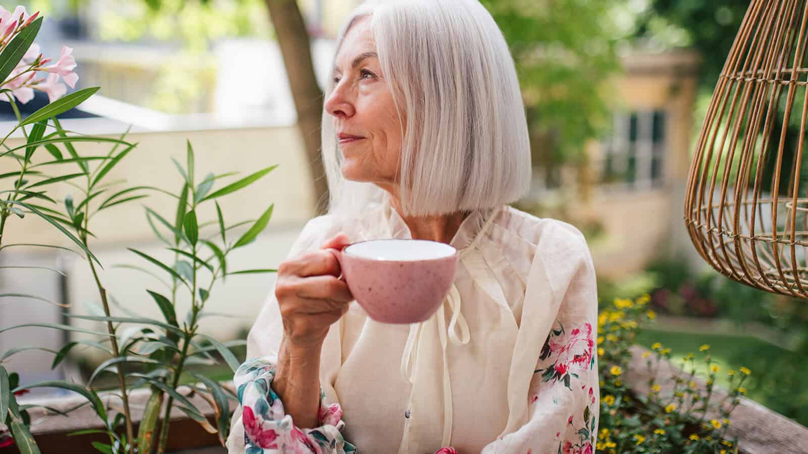 An older woman with shoulder-length grey hair holds a pink mug while looking out over a garden terrace.