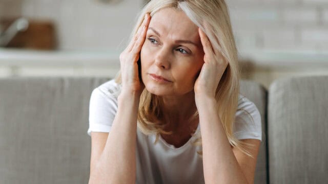 A woman sits with her hands pressed against her temples, appearing stressed or in pain.
