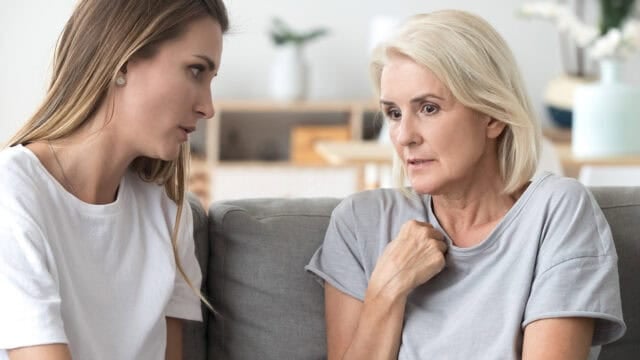 A younger woman sits on a sofa looking concerned while listening to an older woman who is speaking to her.