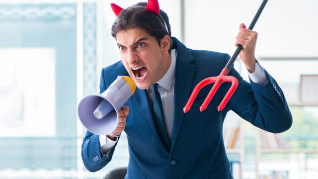 A man in a suit wearing red devil horns shouts into a megaphone while holding a toy pitchfork.