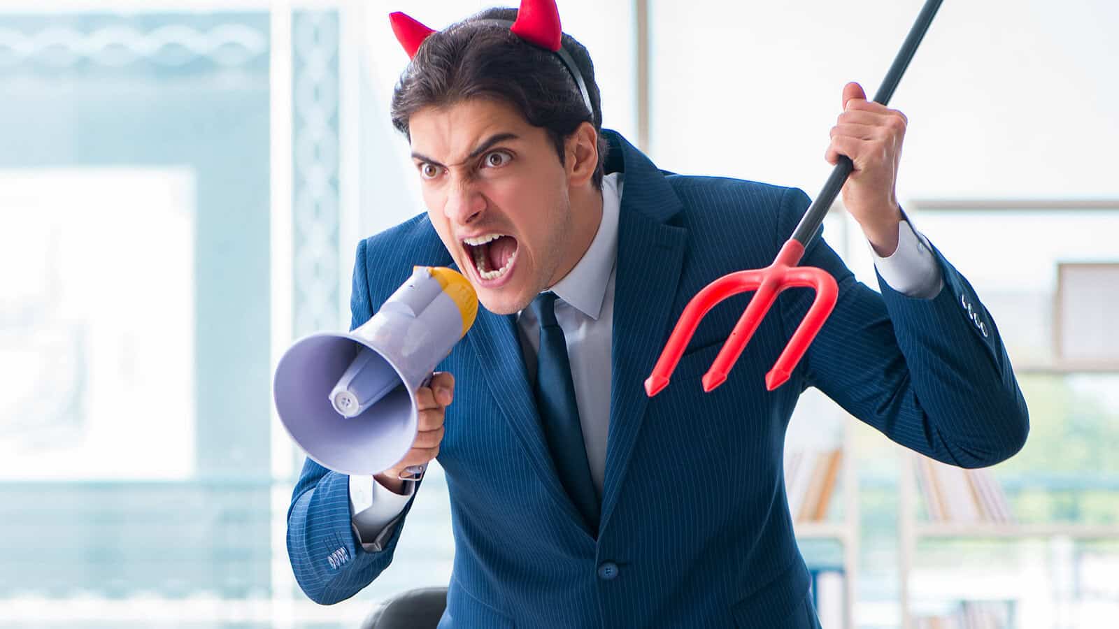 A man in a suit wearing red devil horns shouts into a megaphone while holding a toy pitchfork.