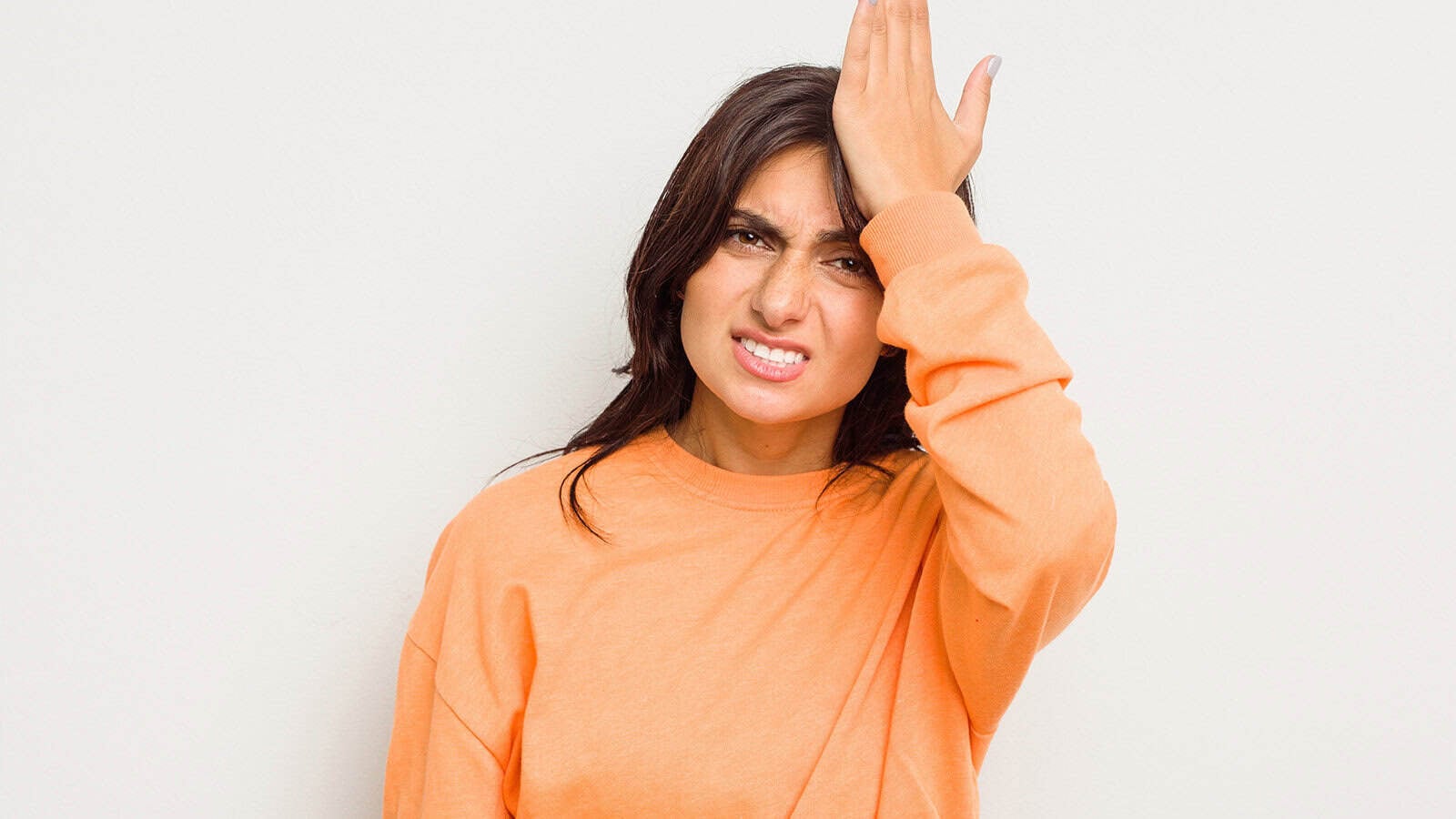 A woman in an orange long-sleeved shirt looking frustrated while slapping her forehead with her hand.