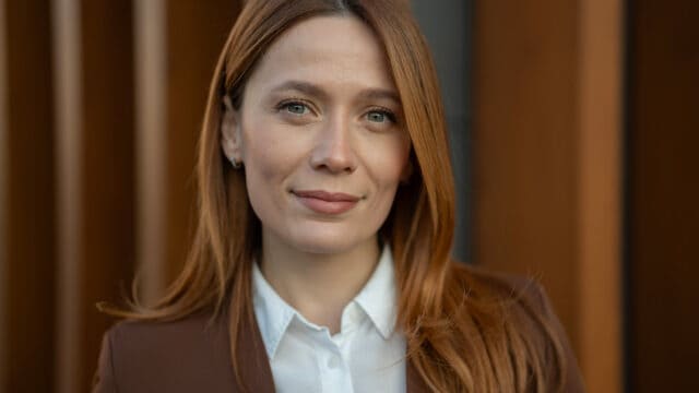 A woman with reddish-brown hair wearing a white collared shirt and a brown blazer looks toward the camera.