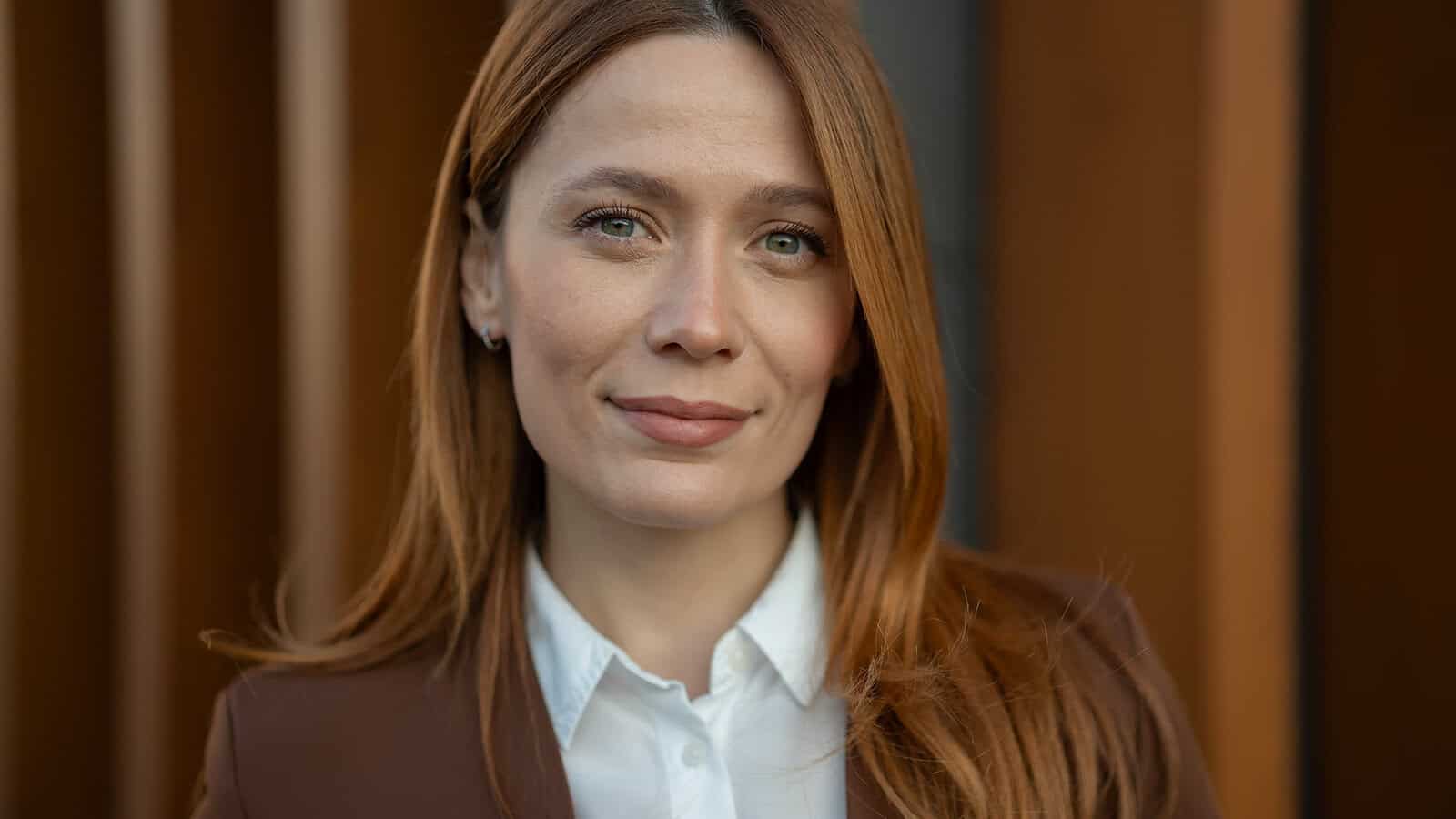 A woman with reddish-brown hair wearing a white collared shirt and a brown blazer looks toward the camera.