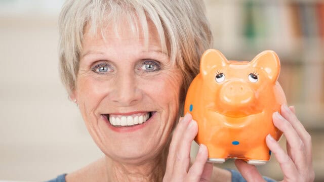 Smiling older woman with short gray hair holds an orange piggy bank close to her face, showing her teeth. The background is softly blurred, focusing on her joyful expression and the piggy bank.