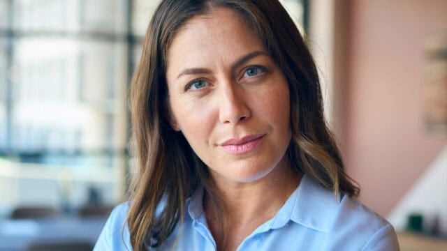 A woman with long brown hair wearing a light blue collared shirt looks directly at the camera.